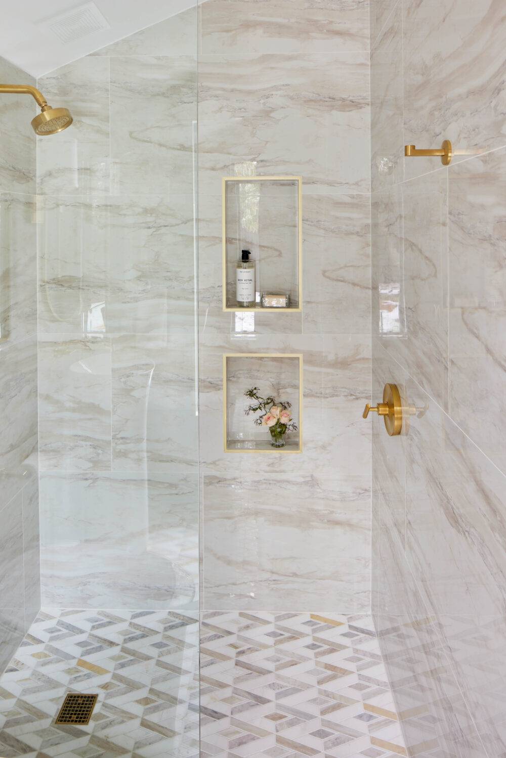 Shower with cream-colored marble tiled walls and white and taupe mosaic tile floor. 