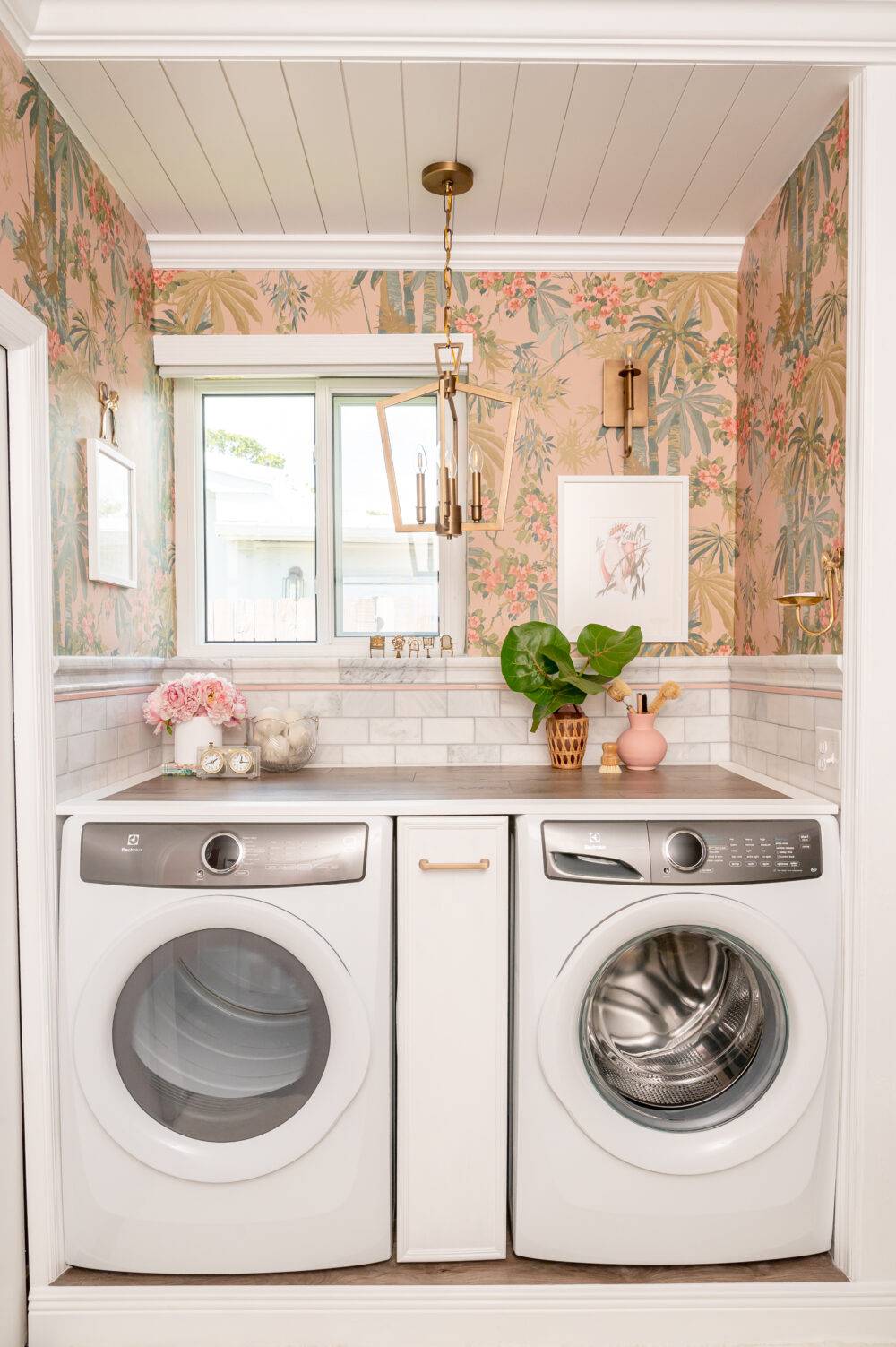 Washer and dryer with small plants and nicknacks. Bright pink floral wallpaper and marble tile.
