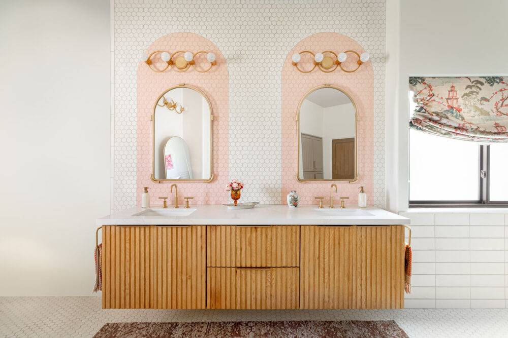 Double sink vanity with mirrors, gold light fixtures and white with pink arch tile.