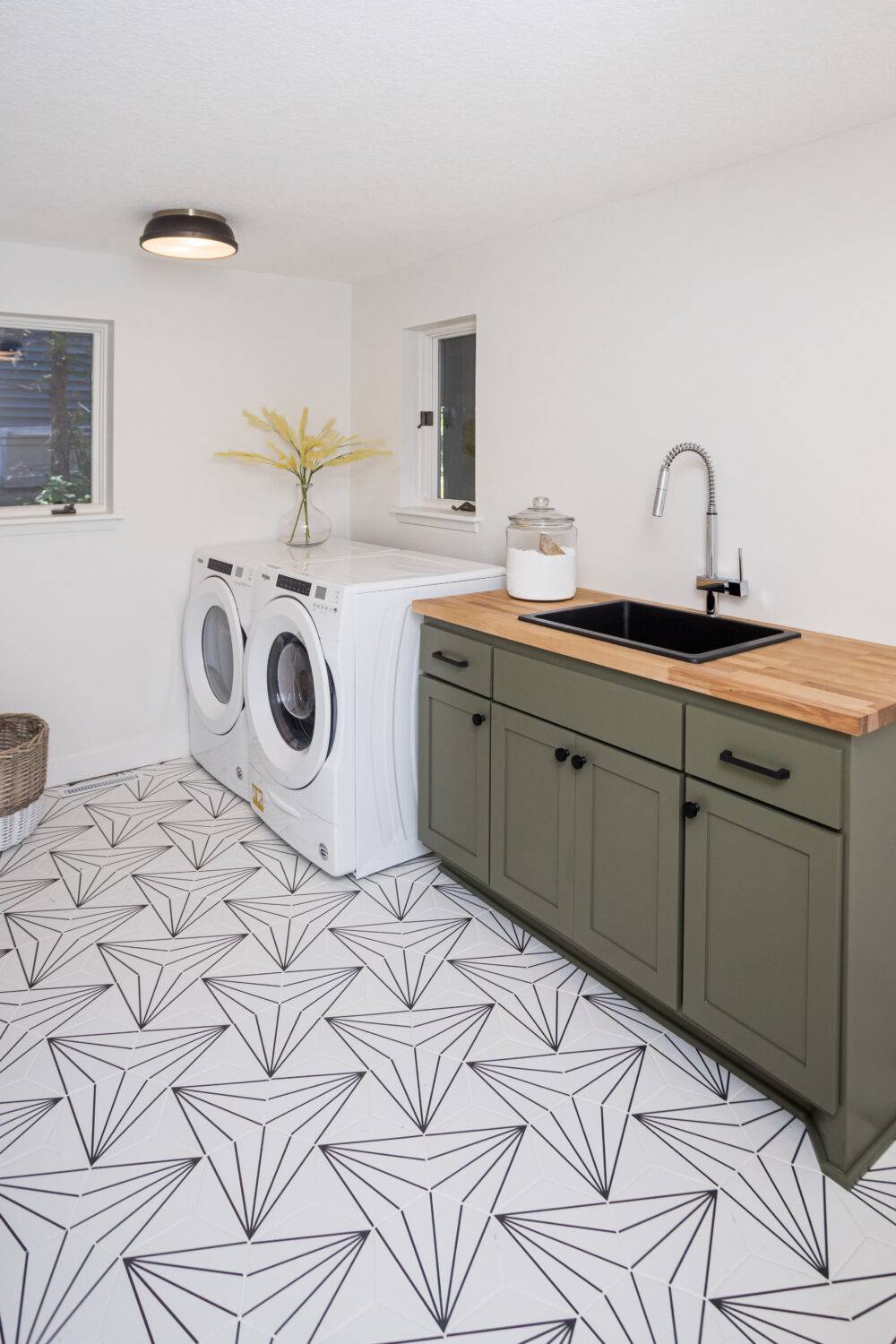 White laundry room with olive green cabinets and black geometric patterned floor tile.
