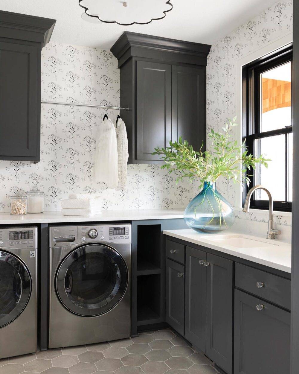 Laundry room with delicate grey and white floral wallpaper. Large blue vase with green-leaved twigs. Grey hexagon tile floor. 